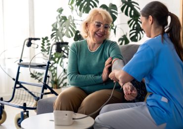Beautiful senior woman measuring blood pressure with help of home nurse, sitting on sofa in living room, feeling satisfied and happy with home care. Female nurse assisting senior woman to measure blood pressure